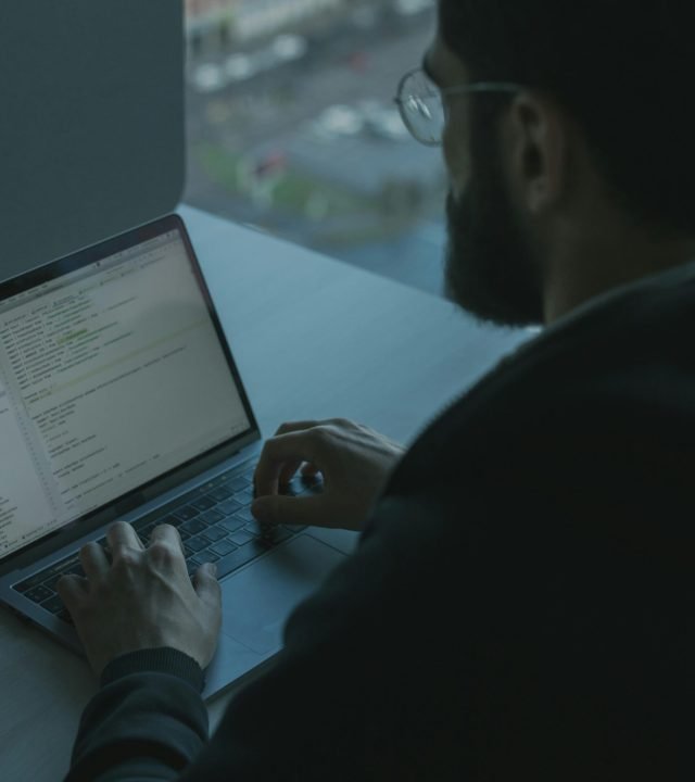 A man coding on his laptop by a window in an office setting, showcasing technology work.